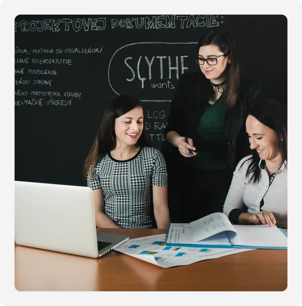 There are three girls in front of the blackboard, one standing and two sitting at the side of the table.
