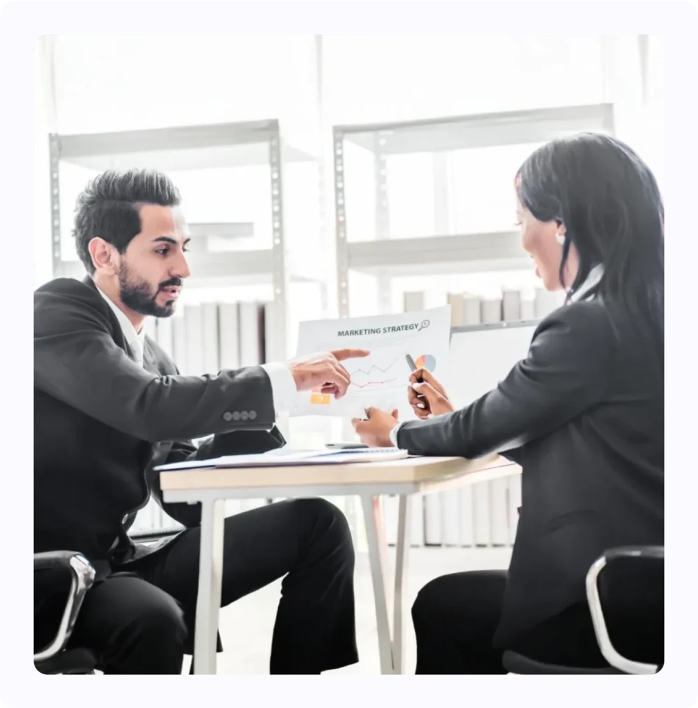 Two business professionals in formal attire discussing a marketing strategy chart in a modern office setting.