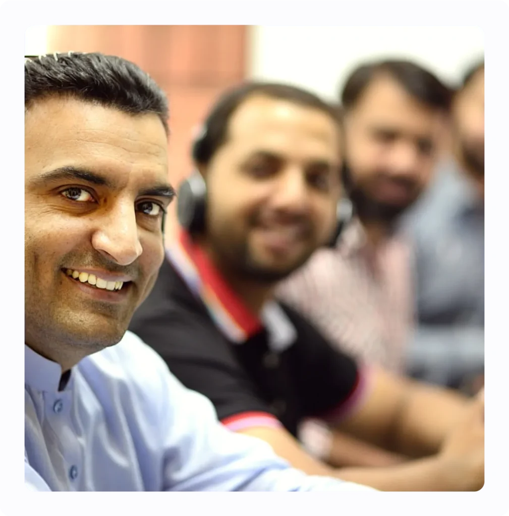 A group of smiling men sitting in a row, with the man in the foreground wearing a light blue shirt and looking directly at the camera, while the others are slightly blurred in the background.